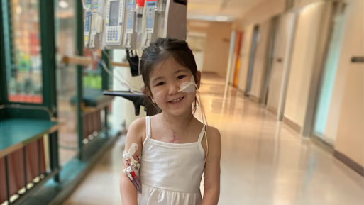 A young girl wearing a white dress smiles as she stands in a hospital corridor, connected to medical equipment. Natural light illuminates the area, and colorful doors are visible in the background.