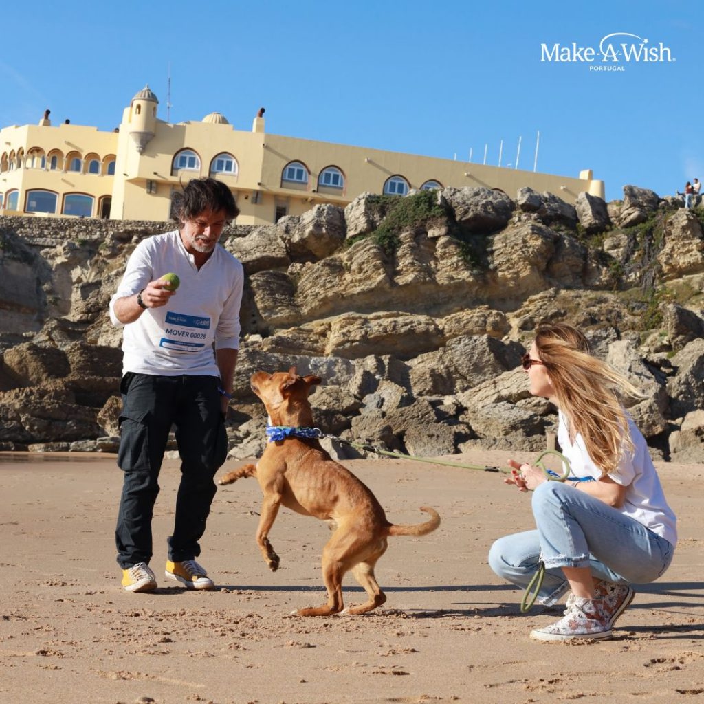 Casal, homem e mulher, com dorsais MOVE FOR WISH KIDS a brincar com o cão na praia