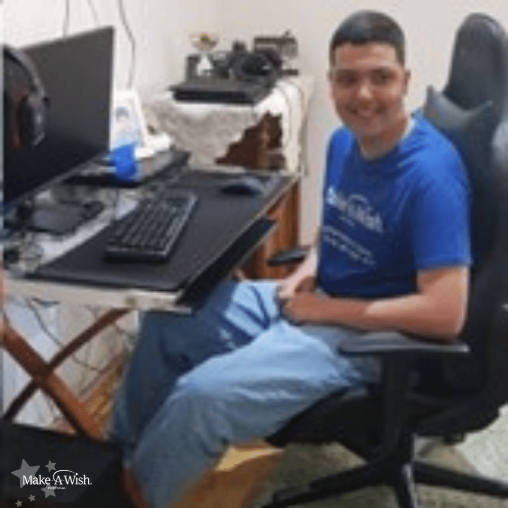 Duarte Silva a sorrir com a t-shirt da Make-A-Wish vestida, sentado na cadeira, à secretária, com o seu novo setup gaming