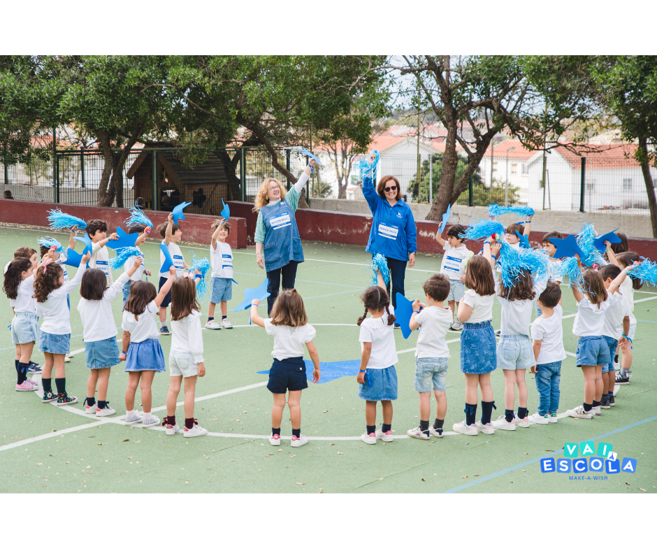 crianças numa escola, com os braços no ar e fitas azuis nas mãos, no exterior, a formarem um círculo à volta de duas professoras vestidas com t-shirts Make-A-Wish