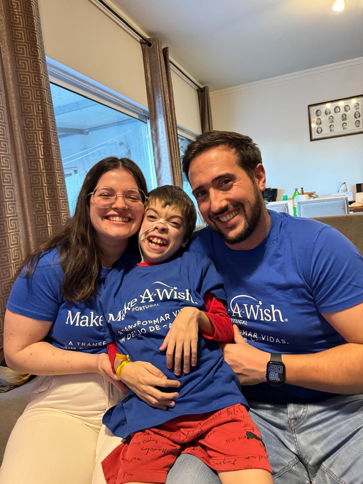 Um rapaz alegre com dois voluntários Make-A-Wish, depois de um desejo realizado. Todos utilizam t-shirts azuis Make-A-Wish.